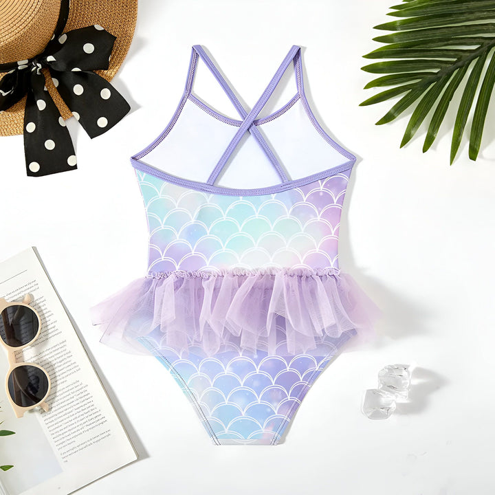 Maillot de bain pour enfant avec motif écailles de sirène et jupon en tulle lilas, chapeau de paille à pois, lunettes de soleil et feuilles de palmier en arrière-plan. Tenue de plage enfantine et colorée.