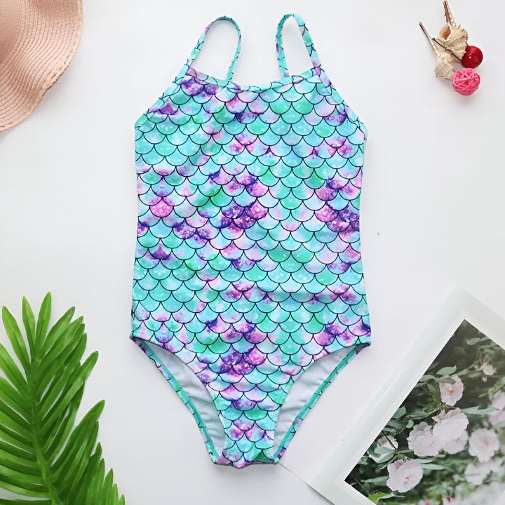 Maillot de bain une pièce pour enfant avec motif écailles de sirène multicolores, chapeau de paille, coquillages et décorations florales autour. Style estival et mignon pour la plage ou la piscine.