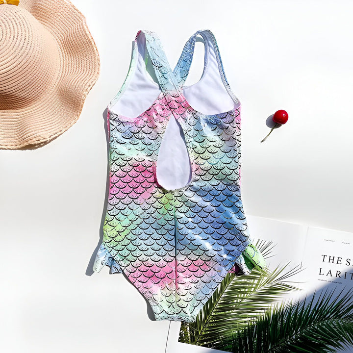 Gros plan d'un maillot de bain pour enfant avec un motif d'écailles de sirène bleu et vert irisé avec des détails brillants, agrémenté de volants et de feuilles de palmier tropicales. Idéal pour les vacances, la plage ou les aventures aquatiques.