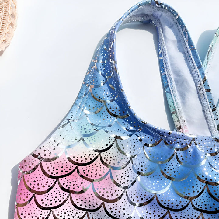 Gros plan d'une queue de sirène aux couleurs arc-en-ciel avec des motifs d'écailles brillantes, et des détails bleus avec des froufrous. En arrière-plan, une feuille de palmier ajoute une touche tropicale.