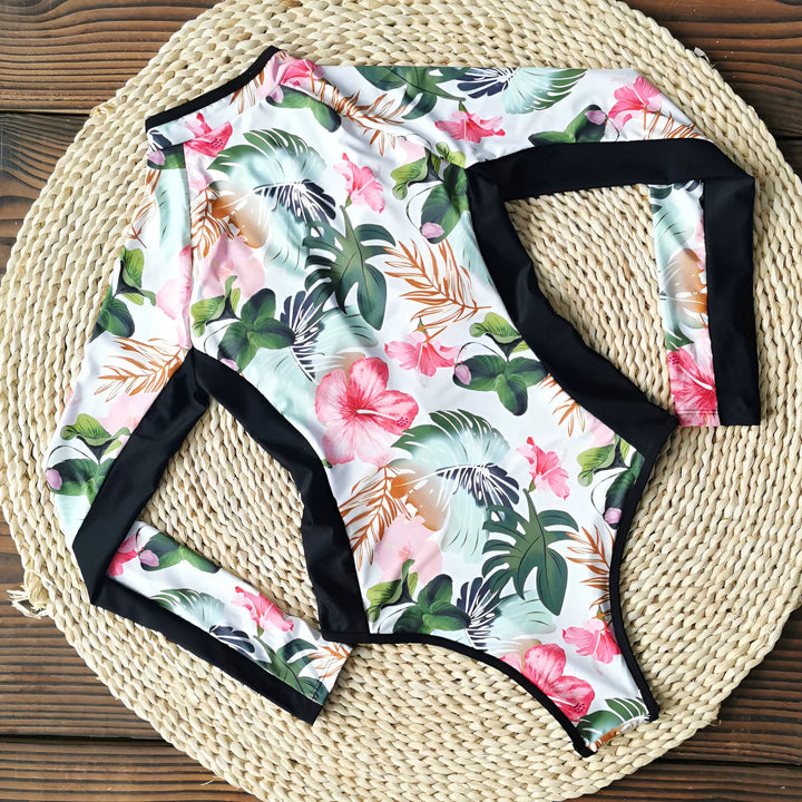 Trois surfeuses se préparent à surfer, marchant sur la plage avec leurs planches. La surfeuse au premier plan porte une combinaison de surf à fleurs. Cheveux longs et blonds. Ambiance décontractée et estivale.