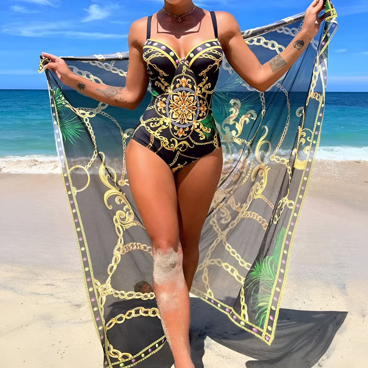 Une femme sur la plage portant un maillot de bain orné de motifs dorés et un paréo noir transparent avec des imprimés de chaînes dorées et de feuilles de palmier. L'eau turquoise de l'océan est visible en arrière-plan.