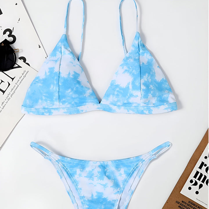 Femme portant un bikini bleu et blanc nuageux, avec de longs cheveux blonds ondulés sur le côté, posant devant un mur blanc. Mode estivale, maillot de bain tendance, look décontracté de vacances.