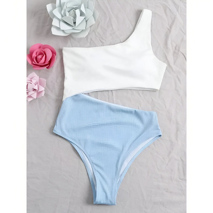 Une femme en maillot de bain deux pièces asymétrique rose et bordeaux, portant un collier carte d'Afrique et une montre. Elle pose sur un pouf tressé.