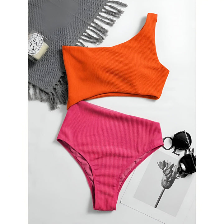 Vue plongeante d'une femme portant un maillot de bain une pièce rose et vert, un chapeau de paille et une veste en jean. Elle est allongée sur un lit. Bijoux aux poignets et tatouages visibles.