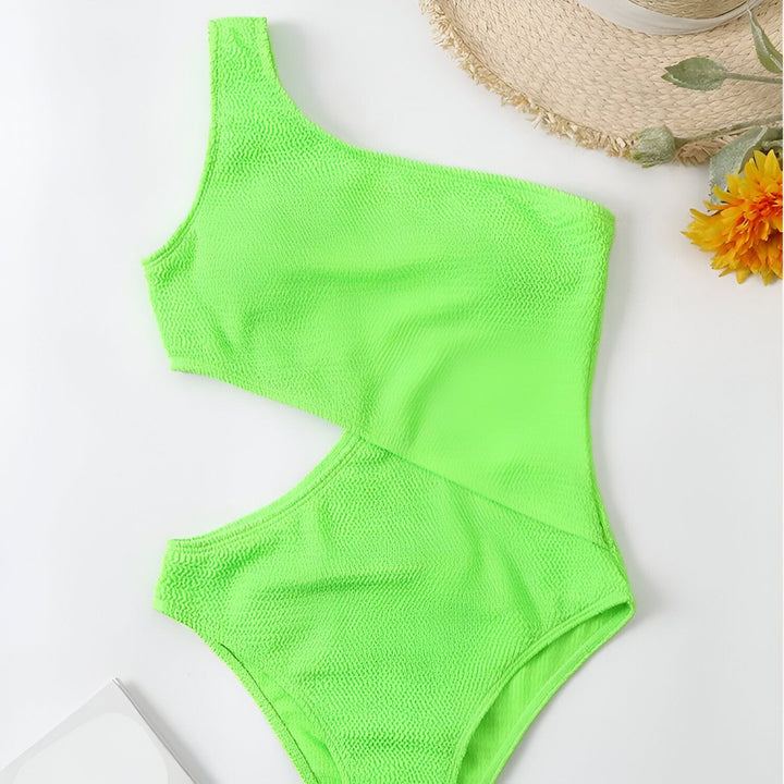 Maillot de bain noir une pièce asymétrique avec découpe sur le côté, accessoirisé d'un chapeau de paille et d'une fleur.