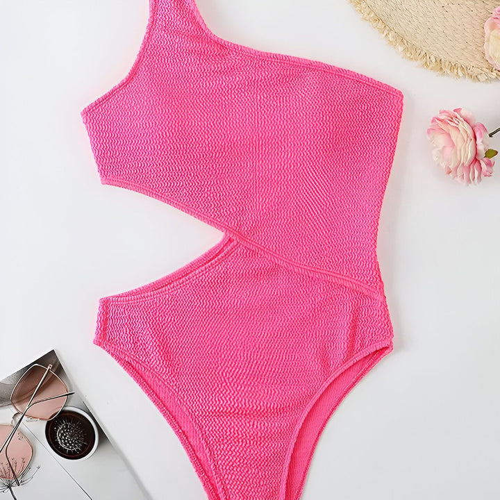 Image d'une femme portant un maillot de bain rose vif une pièce avec une épaule dénudée et une découpe sur le côté, posant sur une serviette blanche à côté d'un livre jaune.