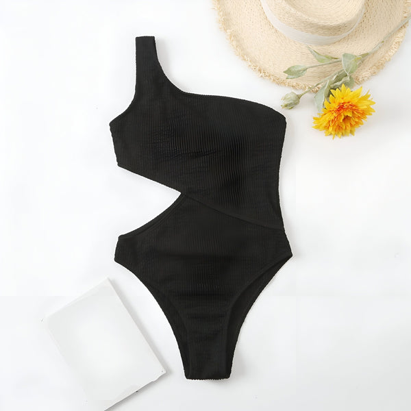 Une femme porte un maillot de bain une pièce noir à une seule épaule avec une découpe sur le côté. Le maillot de bain est texturé et la femme porte un bracelet.
