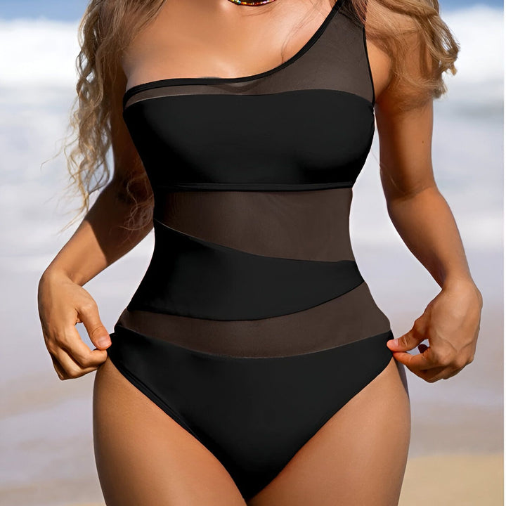 Image d'une femme portant un maillot de bain une pièce noir avec des empiècements en mesh, un design asymétrique avec une seule bretelle. Elle porte un collier coloré. L'arrière-plan est une plage ensoleillée avec un ciel bleu et une mer turquoise.