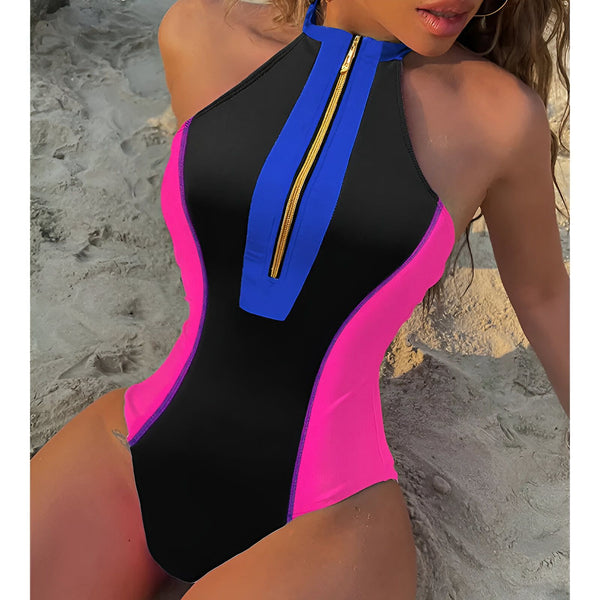 Image rapprochée d'une femme portant un maillot de bain une pièce noir, rose vif et bleu avec une fermeture éclair dorée sur le devant. Le maillot est sans manches et a un col montant. La femme est assise sur le sable.