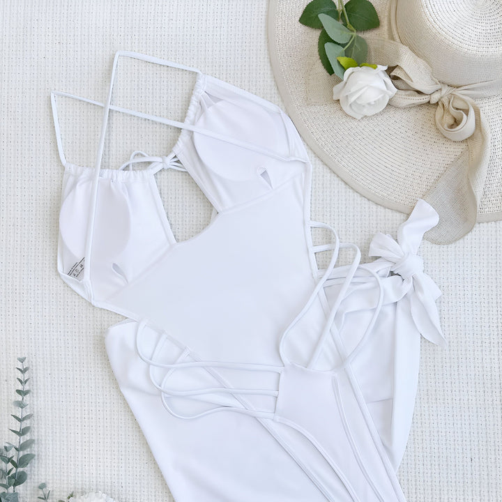 Image d'une femme portant un maillot de bain une pièce blanc avec des découpes originales sur le côté. Le maillot a des bretelles fines et un cordon de serrage au niveau de la poitrine. La femme pose devant un mur de pierres.