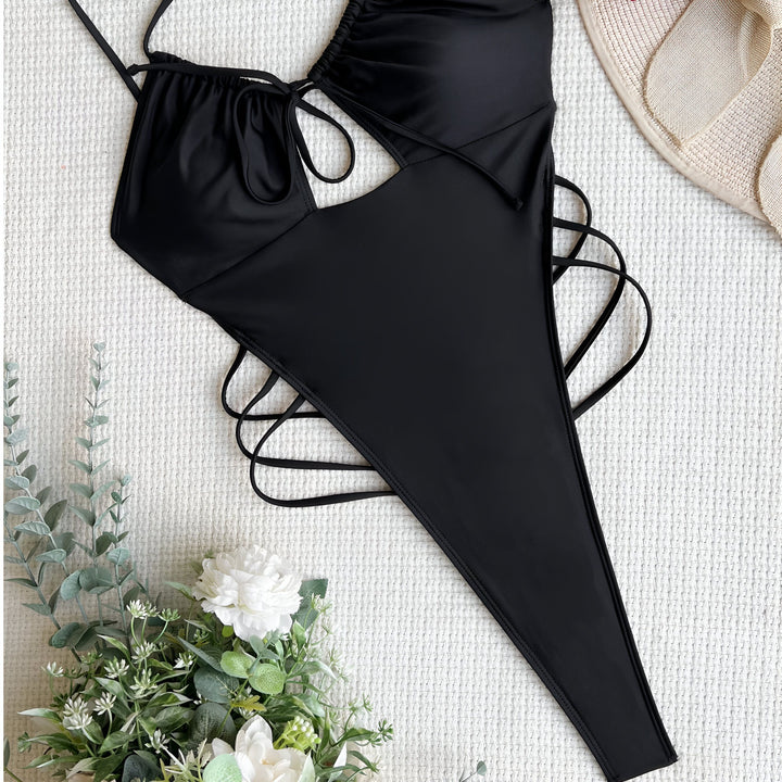 Une femme porte un maillot de bain une pièce blanc avec des détails noués sur le devant et les côtés, et un paréo blanc noué autour de sa taille, devant un mur de pierres claires.