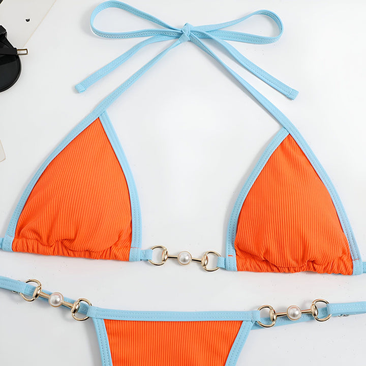 Femme en bikini vert et bleu sur plage de sable blanc, mer turquoise et ciel bleu en arrière-plan. Le bikini est orné de chaînes dorées et de perles. Mode estivale, vacances à la plage.
