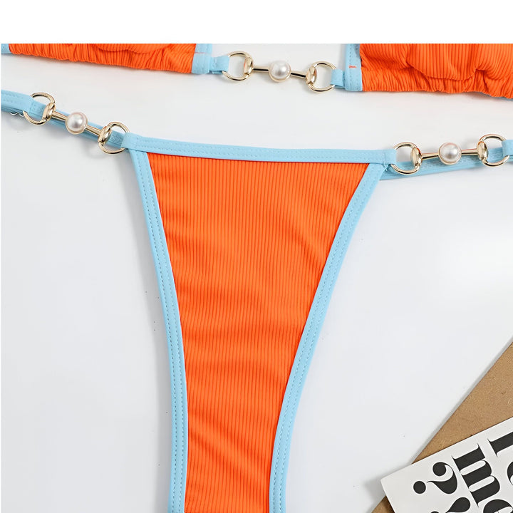 Une femme en bikini jaune fluo et bordures bleues sur une plage ensoleillée. L'eau turquoise et le ciel bleu complètent le tableau. La chaîne de son bikini est ornée de petites perles.