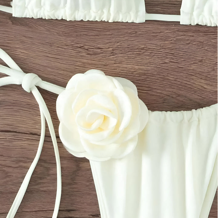 Vue rapprochée d'un maillot de bain blanc deux pièces avec des détails de fleurs blanches, un chapeau de paille, des lunettes de soleil roses et un livre sur une table en bois. Vêtements d'été, mode plage.