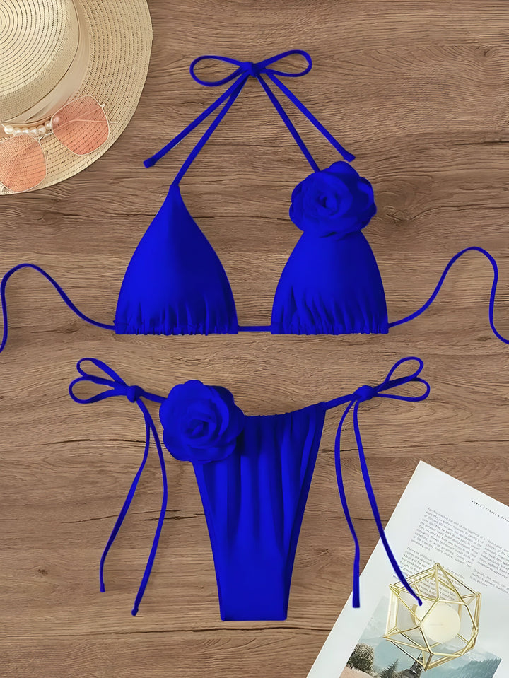 Haut de maillot de bain triangle bleu vif avec des détails floraux, accompagné d'un chapeau de paille et de lunettes de soleil sur une table en bois. Mode estivale.