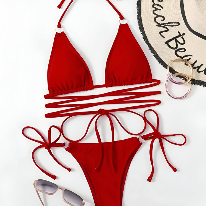 Une femme bronzée porte un bikini rouge vif avec des anneaux métalliques et un collier représentant le continent africain. Le bikini a des liens qui s'enroulent autour de sa taille, mettant en valeur sa silhouette.