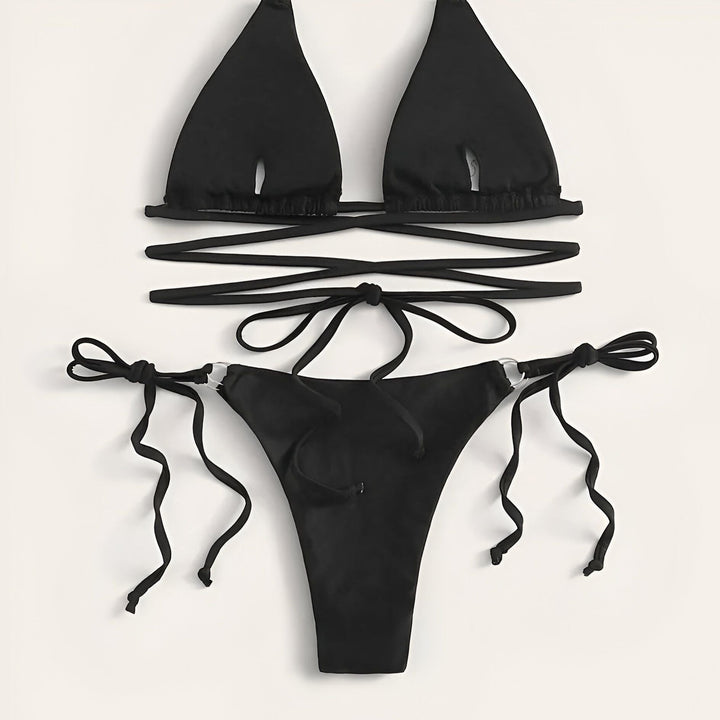 Une femme portant un bikini vert émeraude avec des détails en forme d'anneau et un collier avec un pendentif en forme de carte d'Afrique. Le bikini a des liens croisés autour de sa taille. Elle est assise sur un canapé texturé.