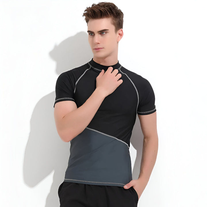Un homme porte un rashguard bleu et marine, debout devant un mur blanc. Vêtements de sport, protection solaire, mode masculine.