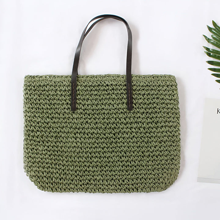 Sac de plage en paille tressée avec anses en cuir noir, style bohème chic. Accessoire de mode estival idéal pour les vacances. Feuille décorative et magazine à proximité.