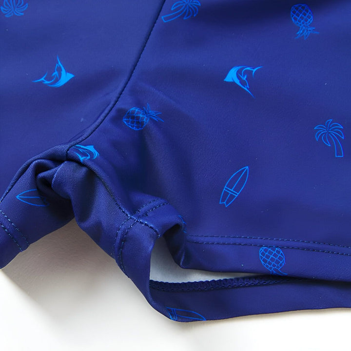 Un short de bain bleu marine pour enfant, avec des motifs d'ananas, de palmiers, de poissons et de planches de surf. Une étoile de mer, une coquille et une feuille tropicale décorent l'ensemble.