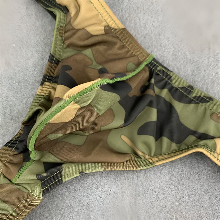 Sous-vêtement masculin de type string avec motif camouflage militaire, sur un fond gris texturé et à côté d'une feuille verte tropicale.