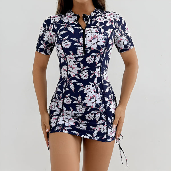 Femme portant un maillot de bain une pièce à fermeture éclair avec un motif floral bleu marine et blanc, manches courtes et taille froncée. Fond blanc. Mode de plage.