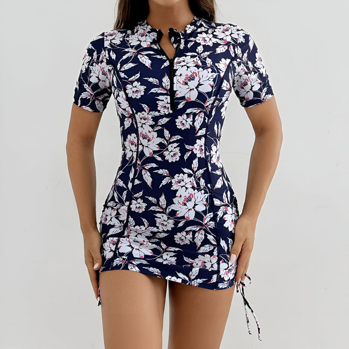 Femme portant un maillot de bain une pièce à fermeture éclair avec un motif floral bleu marine et blanc, manches courtes et taille froncée. Fond blanc. Mode de plage.