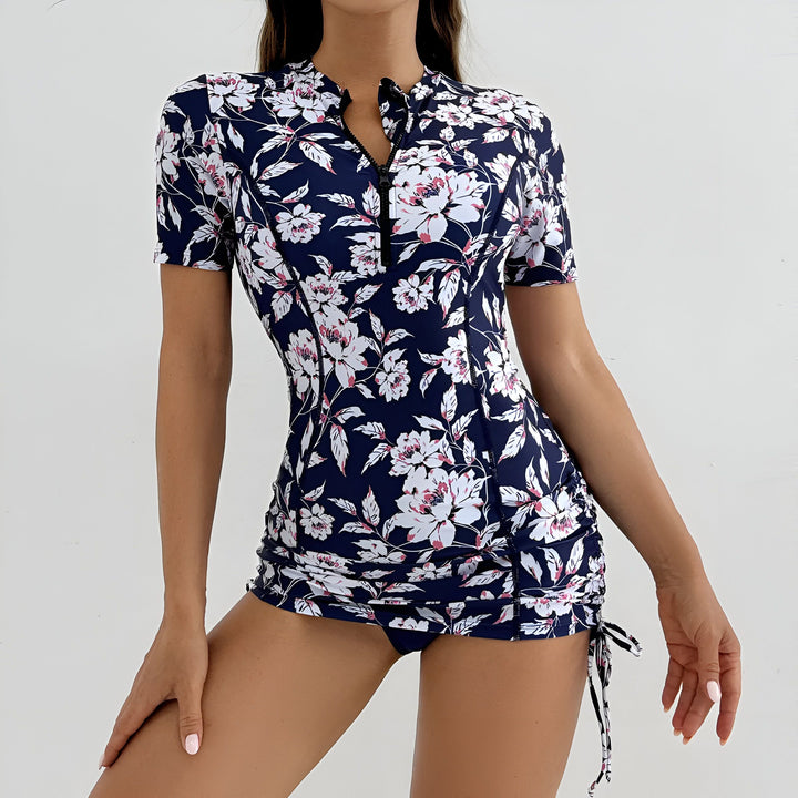 Femme portant une robe de bain une pièce bleue marine à motif floral blanc et rose, avec une fermeture éclair avant et des manches courtes, devant un fond blanc.