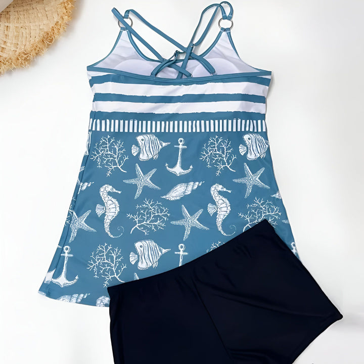 Vue de dessus d'un maillot de bain 2 pièces : une tunique bleue imprimée motifs marins (étoiles de mer, hippocampe, ancre...) et un short de bain noir, avec un chapeau de paille en haut à gauche. Idéal pour la plage ou la piscine.