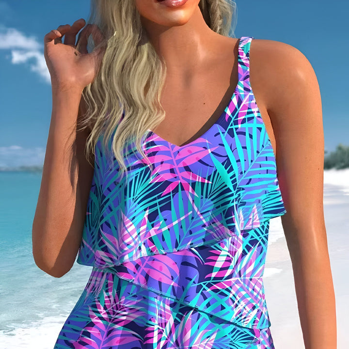 Image d'une femme portant un maillot de bain tankini à motifs tropicaux (bleu, violet, rose) sur une plage ensoleillée. Style décontracté et tendance pour l'été.