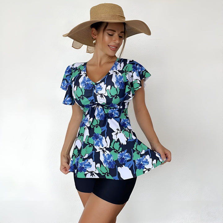 Une femme souriante porte un maillot de bain deux pièces à motif floral bleu et vert avec un chapeau de paille à large bord devant un mur blanc.