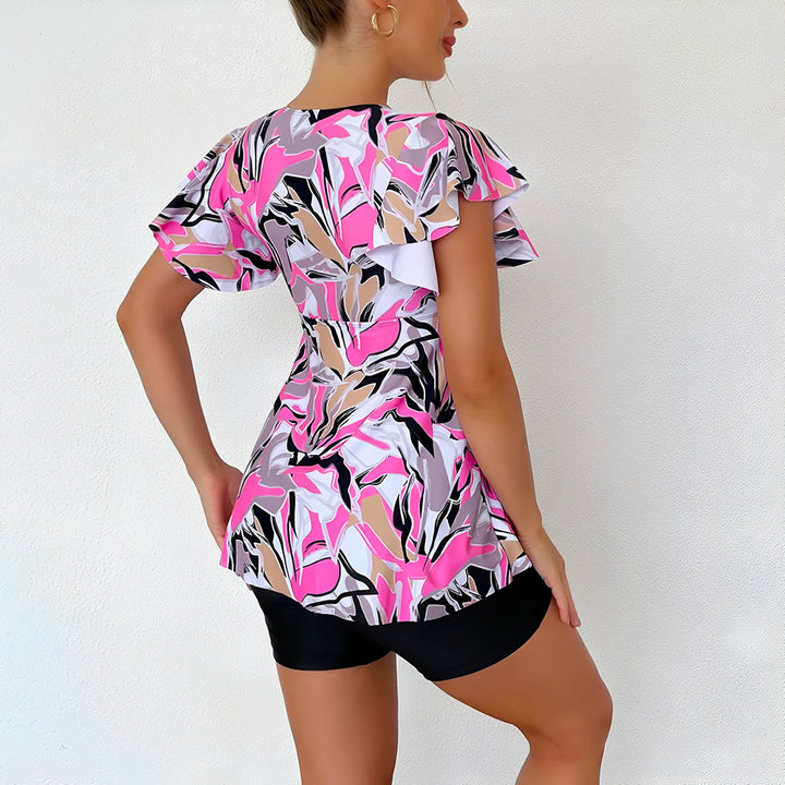 Femme portant un maillot de bain deux pièces avec un haut à motifs rose, noir, beige et blanc, et un bas cycliste noir. Elle pose devant un mur blanc.